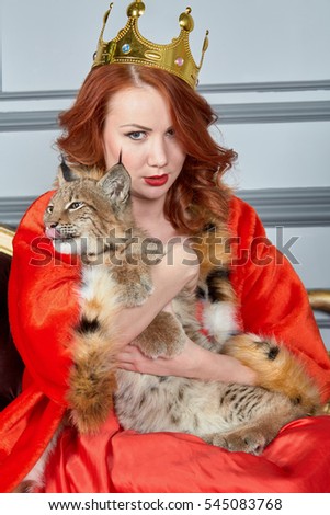 Red-haired woman in red dress, cloak and with crown on head sits on couch holding lynx cub.