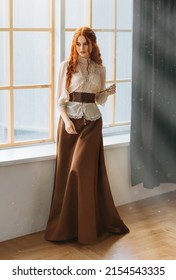 Red-haired woman in vintage dress stands at large classic window waiting love. Clothing costume countess old style white blouse, brown long skirt. Curly red hair. Redhead girl princess 1800s stylish.