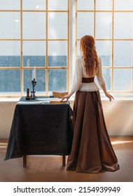 Red-haired woman in vintage dress stands looks at classic window waiting love. Clothing costume countess old style white blouse, brown long skirt. Curly red hair. Redhead girl princess back rear view. - Shutterstock ID 2154939949