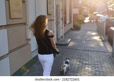 A Red-haired Woman Is Walking With Her Dog On The Sidewalk. A Daily Walk With Your Pet