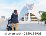 Red-haired woman walks with baby stroller along the touristy promenade of the city of arts and sciences in Valencia, Spain. Palacio de las Artes Reina Sofia building in the background. maternity