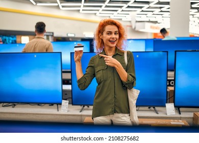 Red-haired Young Woman Chooses Plasma In Store Of Household Appliances, Electronics And Gadgets Holding Credit Card In Her Hand. Buying New TV, Sale Day.