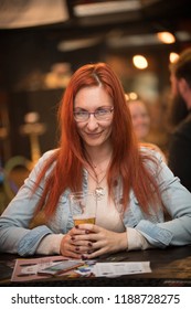 Red-haired young woman in glasses sits in a bar with a mug of beer