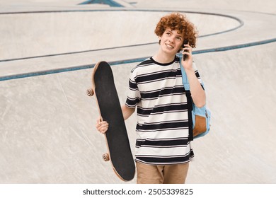 Redhead active young teen boy skater sportsman in casual with bag holds skateboard standing against ramp in city skate park talking on mobile cell smart phone spending time after school lessons - Powered by Shutterstock