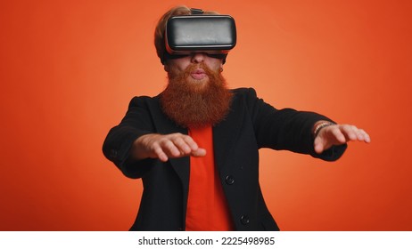 Redhead bearded man using headset helmet app to play simulation innovation game. Watching virtual reality 3D 360 video. Young ginger adult guy boy in VR goggles isolated on orange studio background