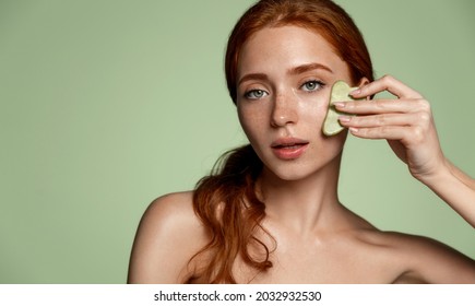 Redhead beautiful woman using skin care acupuncture gua sha jade scraper, face lifting, anti-aging concept. Female model doing cosmetic procedure at home, green background