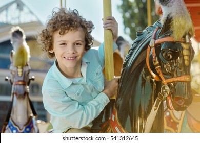 Redhead Boy In The Foreground Of The Camera Rides A Black Horse At The Special Carousel. Healthy And Cheerful Smile Of The Child.