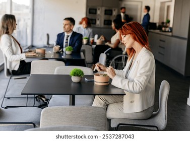 Redhead businesswoman is having a healthy lunch break in the office cafeteria. She is sitting at the table and eating a salad from a takeaway box while her colleagues are talking in the background - Powered by Shutterstock