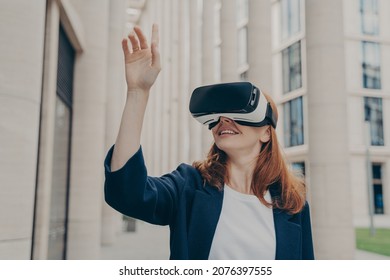 Redhead businesswoman managing business project through virtual reality platform. Smiling young female office worker enjoying spending time with VR headset glasses, pointing with forefinger up in air - Powered by Shutterstock