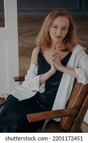 Redhead Confident Woman In Black Evening Dress And White Shirt. Stylish Mid Adult Lady Is Sitting In An Arm Chair. Happy Rich European Woman Is Going Off To A Party. Fashionable Trendy Outfit.