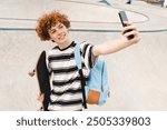 Redhead curly positive high school pupil student teen boy guy teenager skater in casual with backpack holds skateboard smart phone taking selfie shot against in city skate park riding on ramp