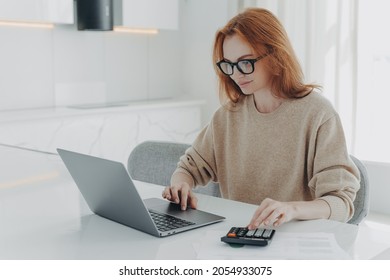 Redhead European Female Accountant Analyzes Documents Uses Calculator Makes Report Uses Laptop Computer Sits At White Desktop Wears Spectacles Beige Jumper Poses Against White Home Interior.