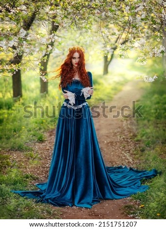 Redhead fantasy woman queen. Blue long velvet medieval dress, vintage clothing. Red curly hair flying waving in wind. Summer nature green flowering trees garden footpath path. Girl Princess walking.