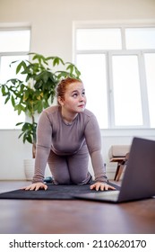 Redhead Fat Woman Standing With Knees On Fitness Mat, Ready To Train, At Home In Living Room, Caucasian Overweight Lady In Sportive Outfit Want To Lose Weight, Watch Video Training On Laptop