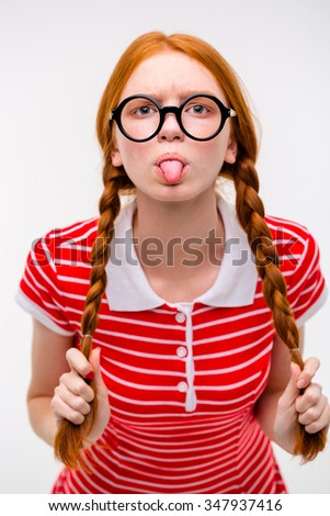 Redhead funny amusing girl with two braids in red striped t-shirt and round glasses showing tongue posing on white background