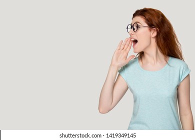 Redhead girl hold hand near mouth shouts speak aloud turn head aside face profile view look at copy space, woman make announcement advertisement freespace for text ad concept image on grey studio wall