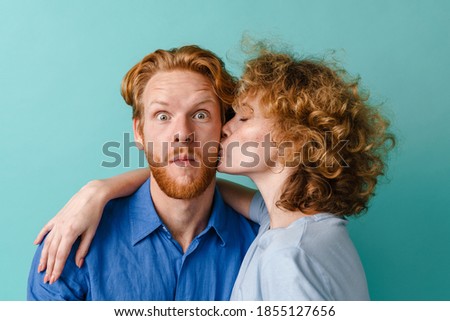 Redhead girl kissing and hugging her surprised boyfriend isolated over blue background