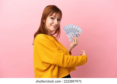 Redhead Girl Taking A Lot Of Money Isolated On Pink Background Pointing Back