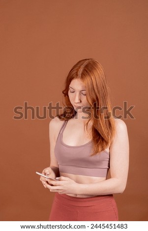 A redhead girl, in a vertical composition against a brown background, gazes directly at her mobile phone while using it, conveying a sense of connectivity and engagement.