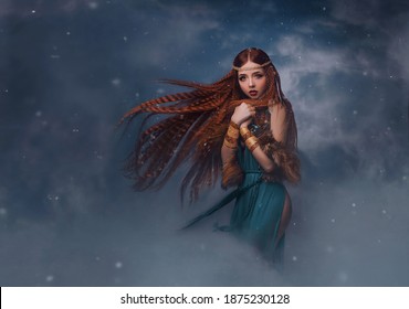 redhead goddess fantasy woman walks in the clouds. Fashion model posing in studio background dramatic winter sky with smoke. Elf princess girl. Long red hair flying in wind snow is falling. Blue dress