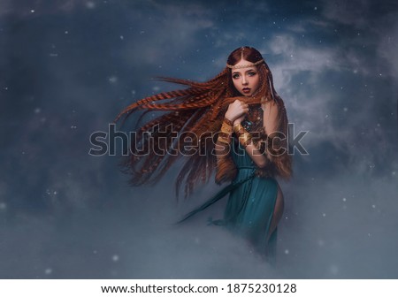 redhead goddess fantasy woman walks in the clouds. Fashion model posing in studio background dramatic winter sky with smoke. Elf princess girl. Long red hair flying in wind snow is falling. Blue dress