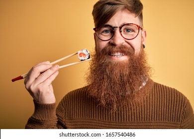 Redhead Irish man with beard eating salmon maki sushi using chopsticks over yellow background with a happy face standing and smiling with a confident smile showing teeth