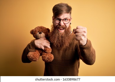 Redhead Irish man with beard hugging teddy bear stuffed animal over yellow isolated background annoyed and frustrated shouting with anger, crazy and yelling with raised hand, anger concept