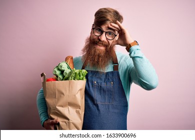 Redhead Irish Supermarket Worker Man With Beard Wearing Apron And Holding Fresh Groceries Stressed With Hand On Head, Shocked With Shame And Surprise Face, Angry And Frustrated. Fear And Upset