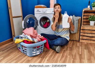 Redhead man with long beard doing laundry holding detergent bottle covering mouth with hand, shocked and afraid for mistake. surprised expression 