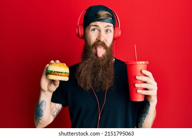 Redhead man with long beard eating a tasty classic burger and drinking soda sticking tongue out happy with funny expression.  - Shutterstock ID 2151795063