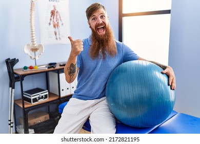Redhead man with long beard holding pilates ball at rehabilitation clinic pointing thumb up to the side smiling happy with open mouth 