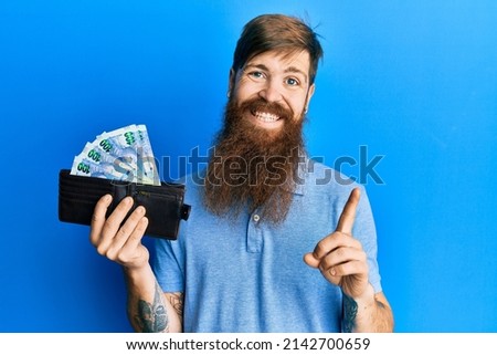Redhead man with long beard holding wallet with south african rand banknotes smiling happy pointing with hand and finger to the side 