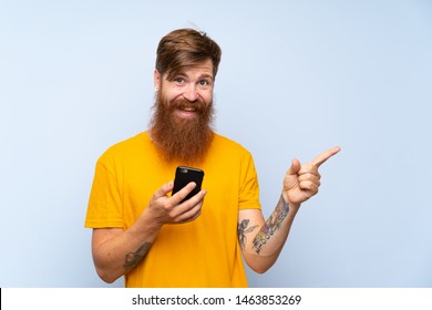 Redhead Man With Long Beard With A Mobile Over Isolated Blue Wall Surprised And Pointing Finger To The Side