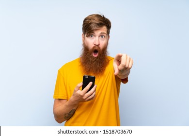 Redhead Man With Long Beard With A Mobile Over Isolated Blue Wall Surprised And Pointing Front