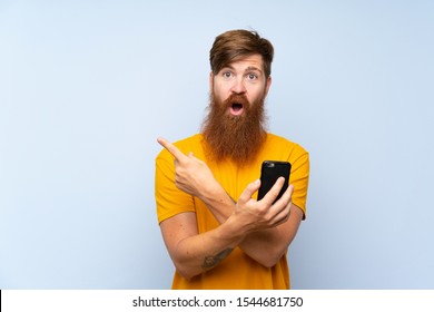 Redhead Man With Long Beard With A Mobile Over Isolated Blue Wall Surprised And Pointing Side