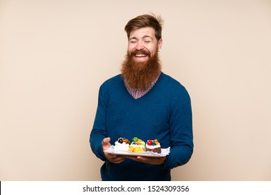 Redhead man with long beard over isolated background holding mini cakes
