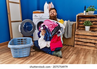 Redhead Man With Long Beard Putting Dirty Laundry Into Washing Machine Puffing Cheeks With Funny Face. Mouth Inflated With Air, Catching Air. 