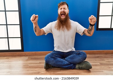Redhead man with long beard sitting on the floor at empty room celebrating mad and crazy for success with arms raised and closed eyes screaming excited. winner concept 