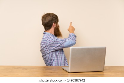 Redhead Man With Long Beard In A Table With A Laptop Pointing Back With The Index Finger