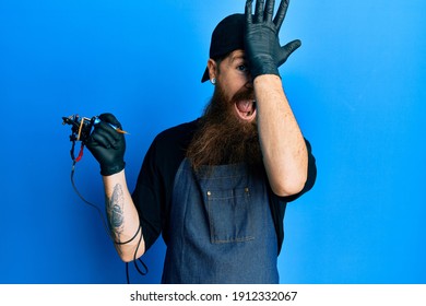 Redhead Man With Long Beard Tattoo Artist Wearing Professional Uniform And Gloves Surprised With Hand On Head For Mistake, Remember Error. Forgot, Bad Memory Concept. 