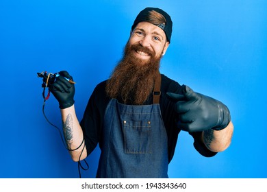 Redhead Man With Long Beard Tattoo Artist Wearing Professional Uniform And Gloves Pointing Fingers To Camera With Happy And Funny Face. Good Energy And Vibes. 