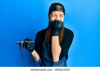 Redhead Man With Long Beard Tattoo Artist Wearing Professional Uniform And Gloves Laughing And Embarrassed Giggle Covering Mouth With Hands, Gossip And Scandal Concept 