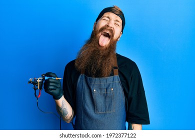 Redhead Man With Long Beard Tattoo Artist Wearing Professional Uniform And Gloves Sticking Tongue Out Happy With Funny Expression. Emotion Concept. 