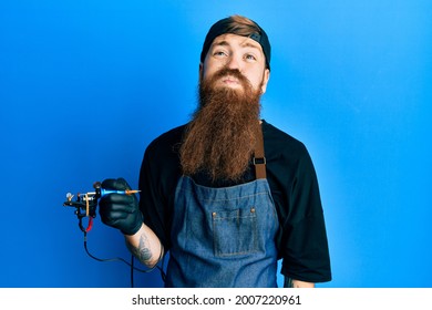 Redhead Man With Long Beard Tattoo Artist Wearing Professional Uniform And Gloves Puffing Cheeks With Funny Face. Mouth Inflated With Air, Crazy Expression. 