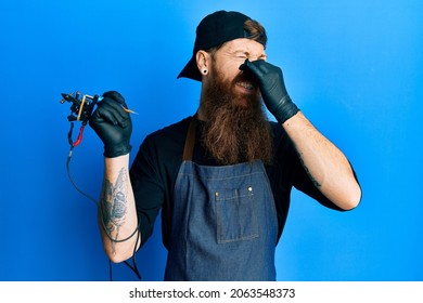 Redhead Man With Long Beard Tattoo Artist Wearing Professional Uniform And Gloves Smelling Something Stinky And Disgusting, Intolerable Smell, Holding Breath With Fingers On Nose. Bad Smell 
