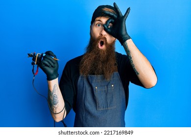 Redhead Man With Long Beard Tattoo Artist Wearing Professional Uniform And Gloves Doing Ok Gesture Shocked With Surprised Face, Eye Looking Through Fingers. Unbelieving Expression. 