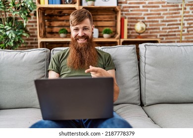 Redhead Man With Long Beard Using Laptop Sitting On The Sofa At The Living Room Smiling Happy Pointing With Hand And Finger 