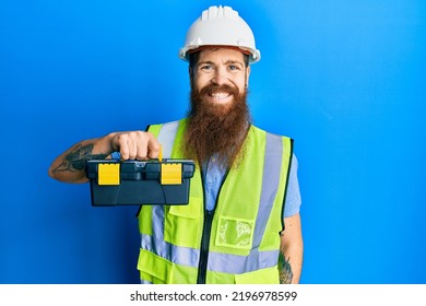 Redhead man with long beard wearing safety helmet and reflective jacket holding toolbox looking positive and happy standing and smiling with a confident smile showing teeth 