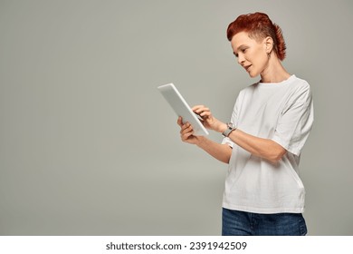 redhead non-binary person in white t-shirt using digital tablet while standing on grey backdrop - Powered by Shutterstock