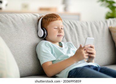 Redhead preteen boy sitting on couch, watching videos or listening to music on mobile phone, using wireless headset, copy space. Modern technologies and kids entertainment concept